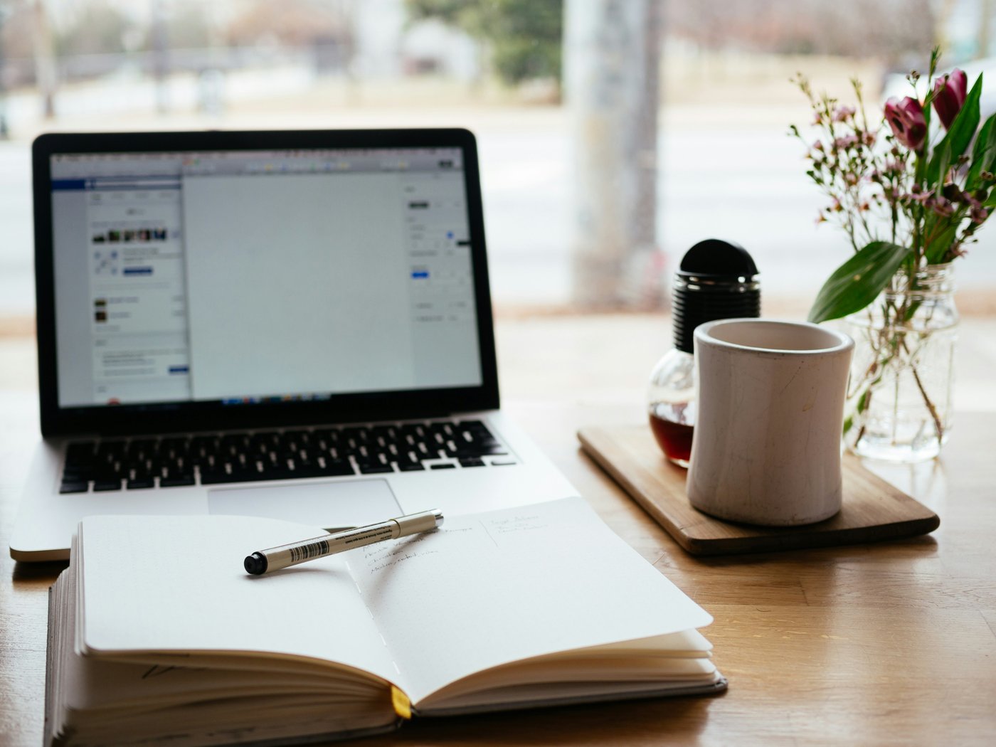 Escritorio con laptop, libreta y café junto a una ventana